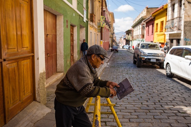 Ein alter Mann steht an einem Gitter gelehnt in einer der Straßen von La Paz. Im Hintergrund sind die bunten Häuser und die Berge zu sehen.